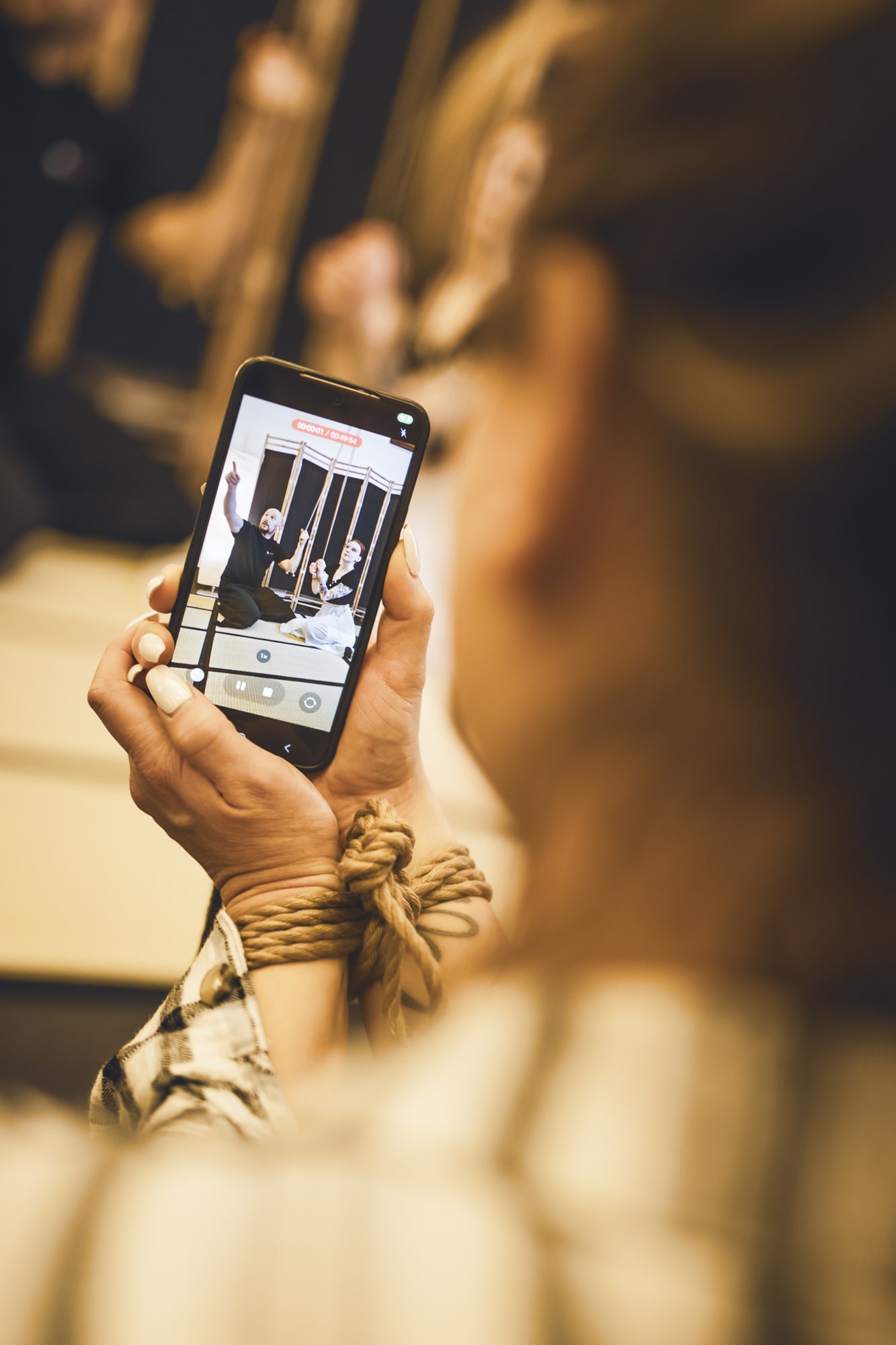 Detail rukou účastnice s provazem kolem zápěstí, která na mobil natáčí Jirku a Myu při ukázce vázání na tatami.