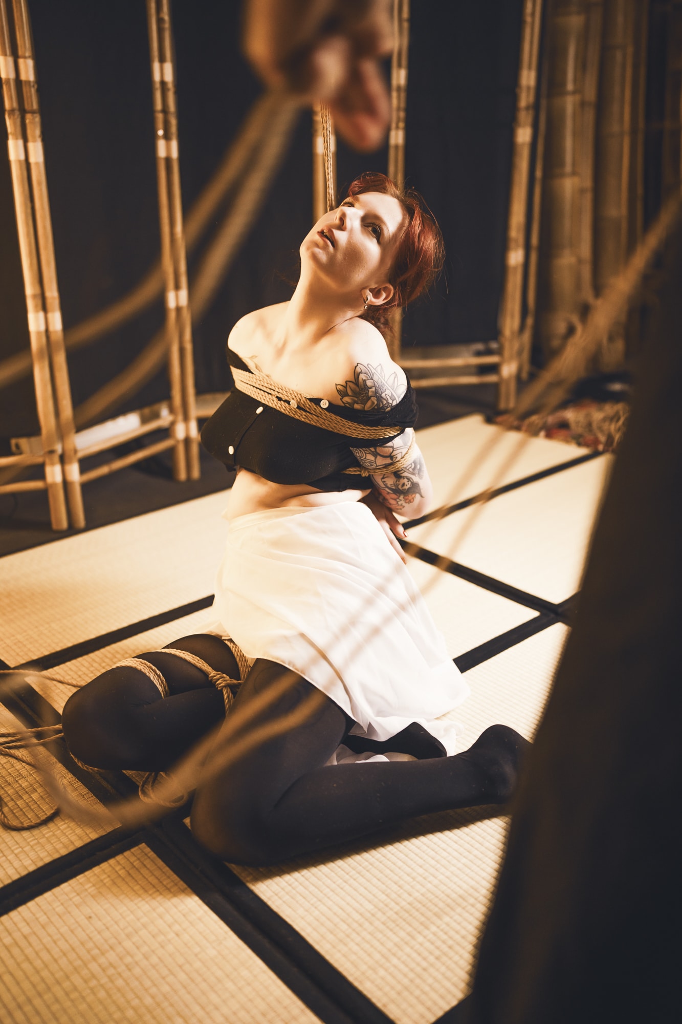 Myu z Alive Kinbaku Dojo klečí na tatami, provazy kolem trupu a nohou, v dojo prostoru s bambusovým pozadím.