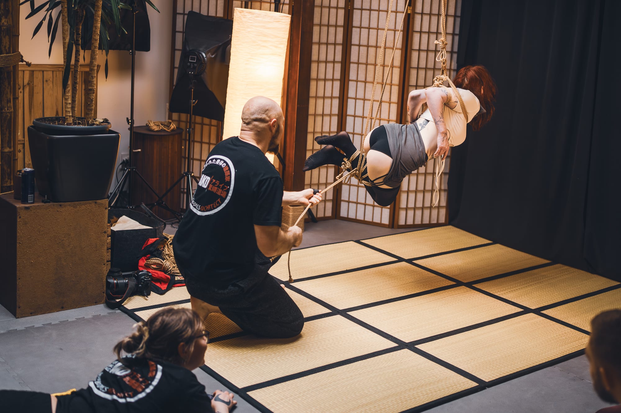 Záběr shora na Myu ležící na tatami při workshopu AKD, s provazem kolem pasu během ukázky techniky.